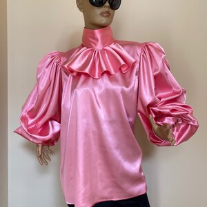 May include: A pink satin blouse with a high ruffled collar and long, puffed sleeves.