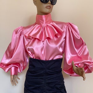 May include: A pink satin blouse with a large ruffled collar and long, puffed sleeves. The blouse is worn with a black skirt.