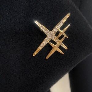 May include: A gold-colored brooch in an abstract starburst design, set with small, sparkling crystals. The brooch is pinned to a black fabric, creating a striking contrast. The design features multiple intersecting lines.