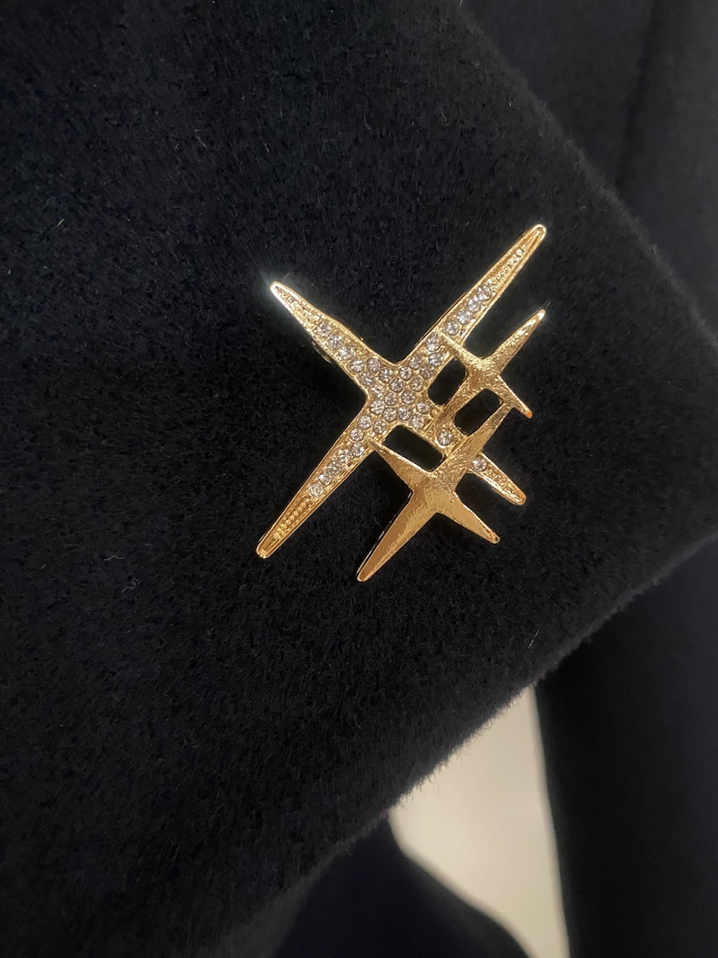 May include: A gold-colored brooch in an abstract starburst design, set with small, sparkling crystals. The brooch is pinned to a black fabric, creating a striking contrast. The design features multiple intersecting lines.
