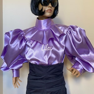 May include: A lilac satin blouse with a high ruffled collar and large puffed sleeves. The blouse has a high neckline and a fitted bodice. The sleeves are long and gathered at the shoulders. The blouse is made of a soft, silky fabric.