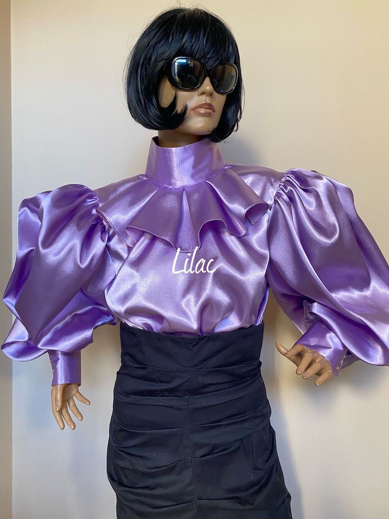 May include: A lilac satin blouse with a high ruffled collar and large puffed sleeves. The blouse has a high neckline and a fitted bodice. The sleeves are long and gathered at the shoulders. The blouse is made of a soft, silky fabric.