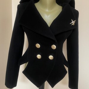 May include: A black double-breasted jacket with a unique design. The jacket features four gold-colored buttons, a wide collar, and a decorative gold-colored brooch. The jacket is displayed on a mannequin.