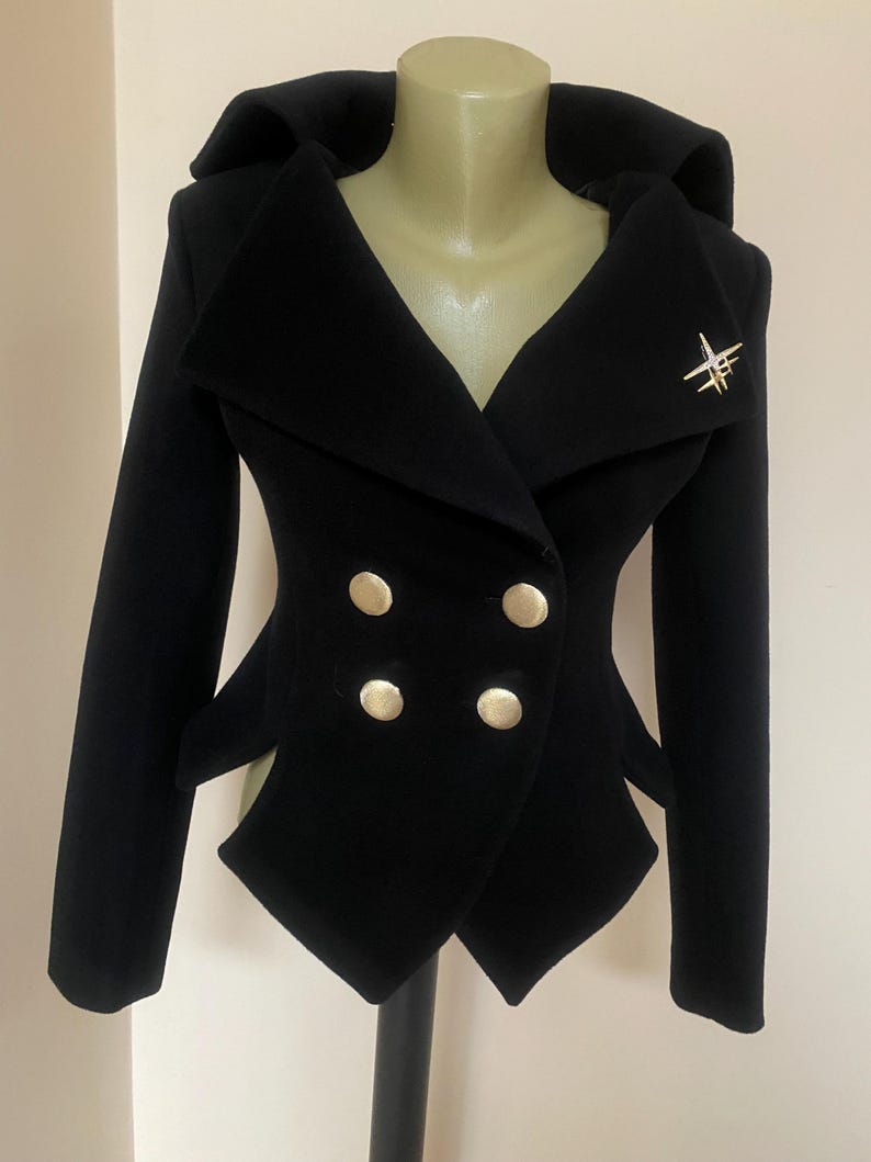 May include: A black double-breasted jacket with a unique design. The jacket features four gold-colored buttons, a wide collar, and a decorative gold-colored brooch. The jacket is displayed on a mannequin.