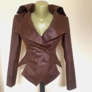 May include: A brown double-breasted jacket with a wide collar and a fitted waist. The jacket has two rows of buttons and unique angular details at the waist. The jacket is displayed on a mannequin.