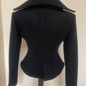 May include: A black jacket with a unique silhouette, featuring a wide collar and long sleeves. The tailored design includes a fitted waist and a vent at the back. The jacket is displayed on a black stand against a neutral background.