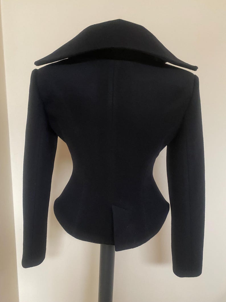 May include: A black jacket with a unique silhouette, featuring a wide collar and long sleeves. The tailored design includes a fitted waist and a vent at the back. The jacket is displayed on a black stand against a neutral background.