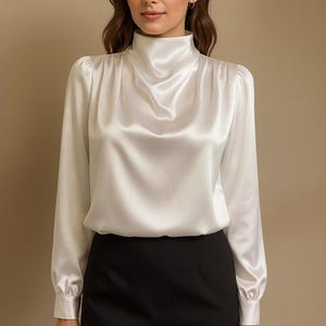 Op de afbeelding: Een witte satijnen blouse met een hoge col en lange mouwen. De blouse wordt gecombineerd met een zwarte rok. De stof heeft een subtiele glans en het ontwerp is klassiek en elegant.