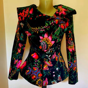 May include: A black jacket with a colourful floral print. The jacket has long sleeves and a distinctive collar design. The floral pattern includes pink, red, yellow, and green flowers and foliage. The waist is fitted.