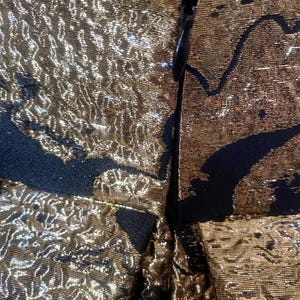 May include: Close-up of a gold and black fabric with a textured, metallic appearance. The fabric is detailed with a black pattern and a zipper closure. The gold color has a shimmering effect.