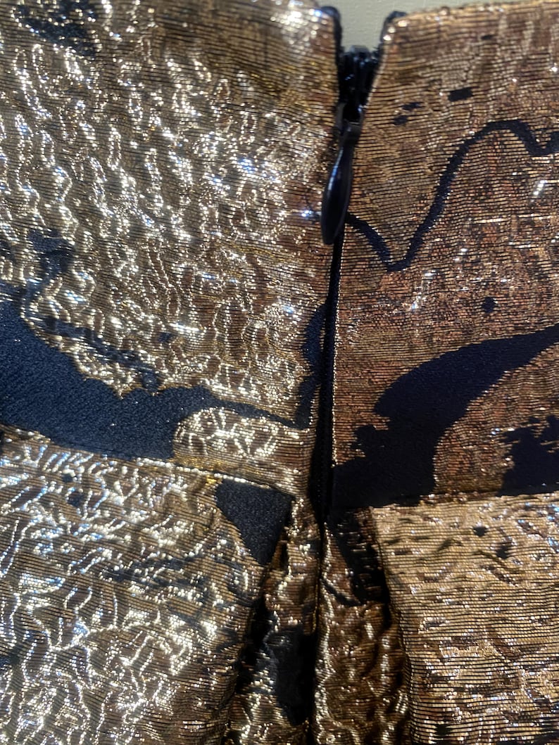 May include: Close-up of a gold and black fabric with a textured, metallic appearance. The fabric is detailed with a black pattern and a zipper closure. The gold color has a shimmering effect.