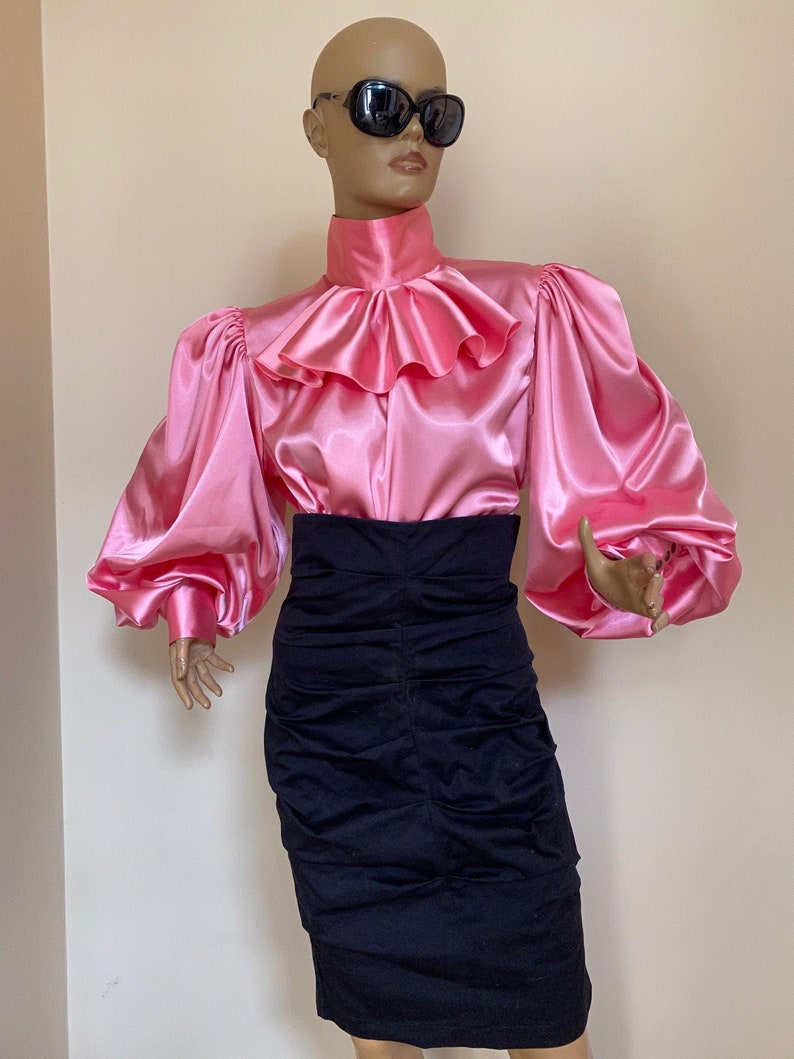 May include: A pink satin blouse with a large ruffled collar and long, puffy sleeves. The blouse is paired with a black skirt.