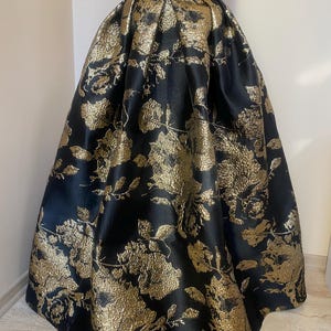 May include: A full-length black skirt with a high waist and a gold floral pattern. The skirt is made of a shiny fabric and has a voluminous silhouette, suitable for formal events.
