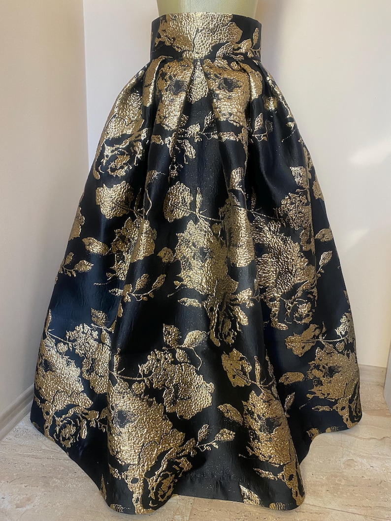 May include: A full-length black skirt with a high waist and a gold floral pattern. The skirt is made of a shiny fabric and has a voluminous silhouette, suitable for formal events.