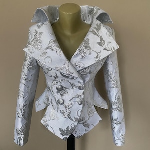 May include: A white jacket with silver floral brocade. The jacket features a wide collar, long sleeves, and a double-breasted closure with silver buttons. The jacket is displayed on a mannequin.