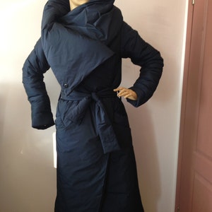 Puede incluir: Un abrigo largo de plumón azul marino con un cuello grande y dramático que se puede llevar arriba o abajo. El abrigo tiene un cinturón y un dobladillo ligeramente asimétrico.