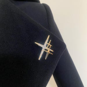 May include: A gold-toned brooch in a starburst design, adorned with clear crystals, is pinned to a dark navy blue coat. The brooch features multiple star shapes, creating a sparkling, eye-catching accessory.