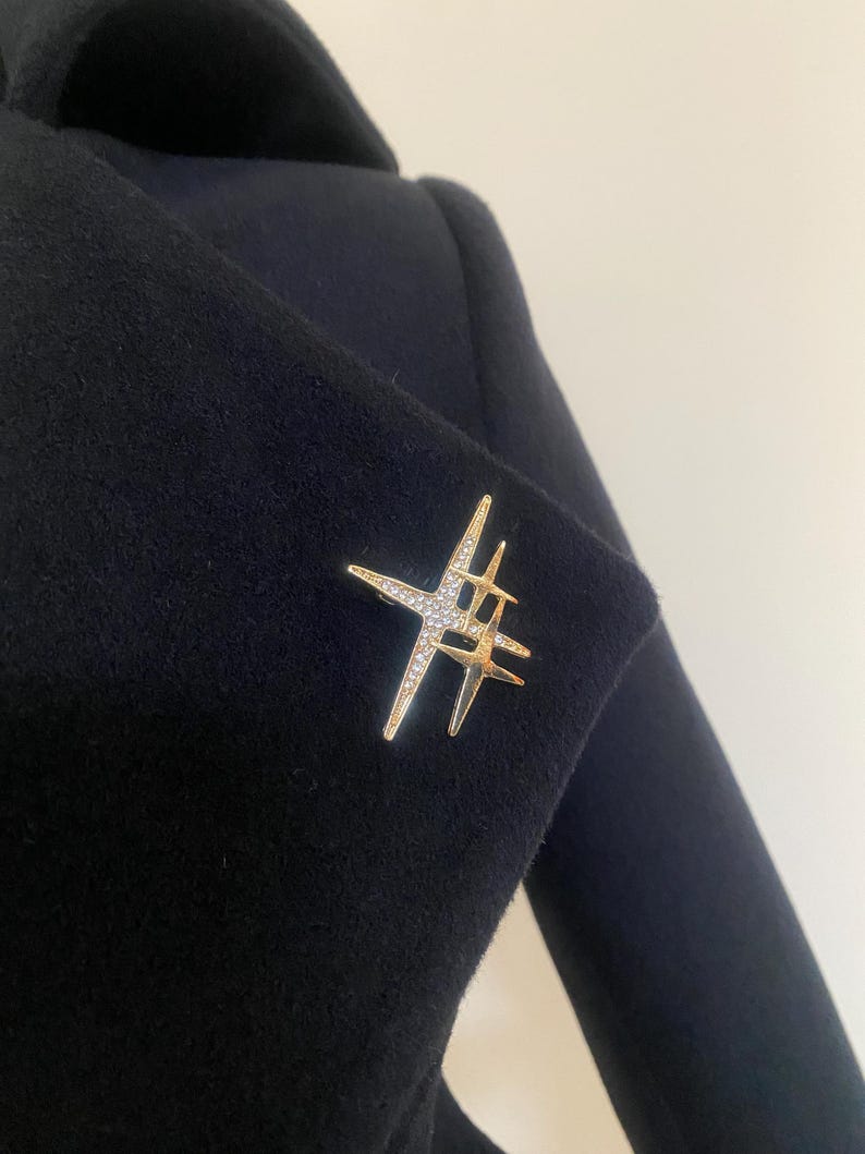 May include: A gold-toned brooch in a starburst design, adorned with clear crystals, is pinned to a dark navy blue coat. The brooch features multiple star shapes, creating a sparkling, eye-catching accessory.