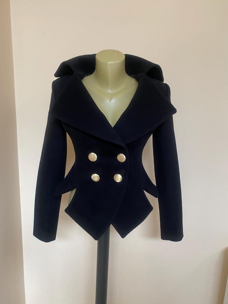 May include: A navy blue double-breasted jacket with a unique design. The jacket features a wide collar, four gold-colored buttons, and distinctive angular cutouts at the waist. The jacket is displayed on a mannequin.