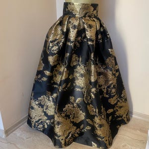 May include: A full-length black skirt with a gold floral pattern. The skirt is displayed on a mannequin and has a high waist and a flared silhouette. The fabric has a metallic sheen, giving it a luxurious appearance.