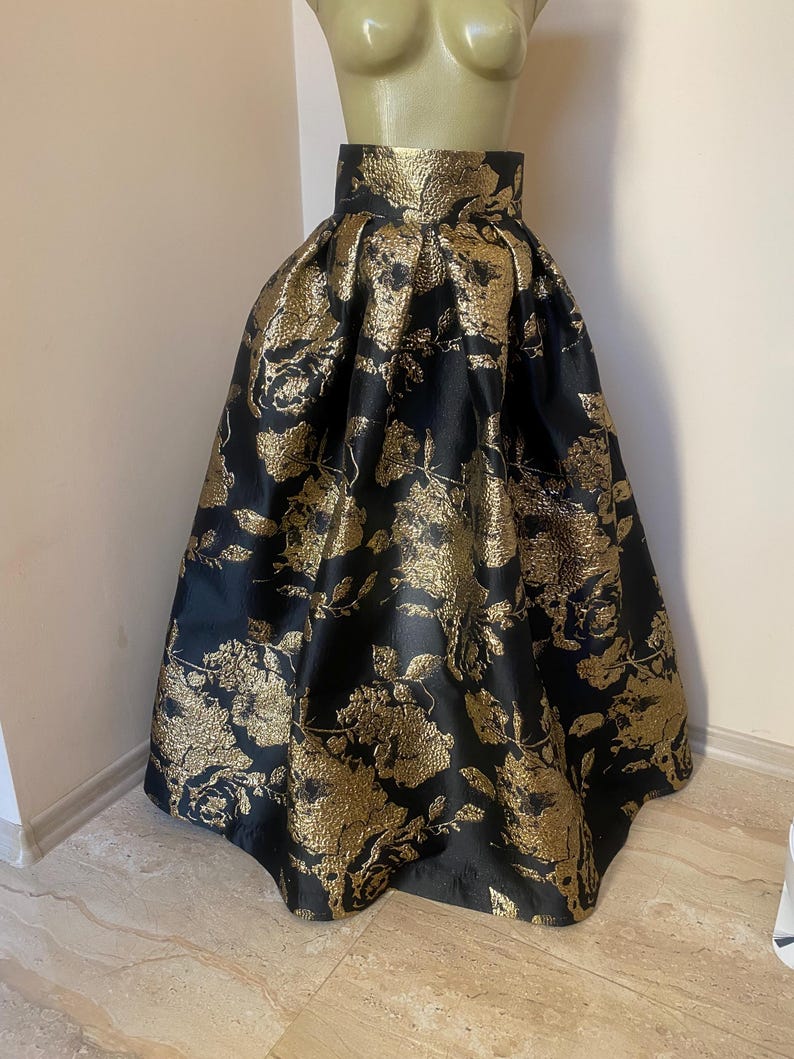 May include: A full-length black skirt with a gold floral pattern. The skirt is displayed on a mannequin and has a high waist and a flared silhouette. The fabric has a metallic sheen, giving it a luxurious appearance.