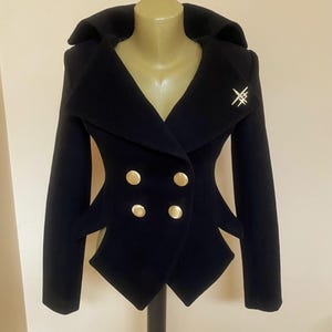 May include: A navy blue double-breasted jacket with gold buttons and a decorative silver pin. The jacket has a unique, tailored design with a wide collar and structured silhouette. The jacket is displayed on a mannequin.