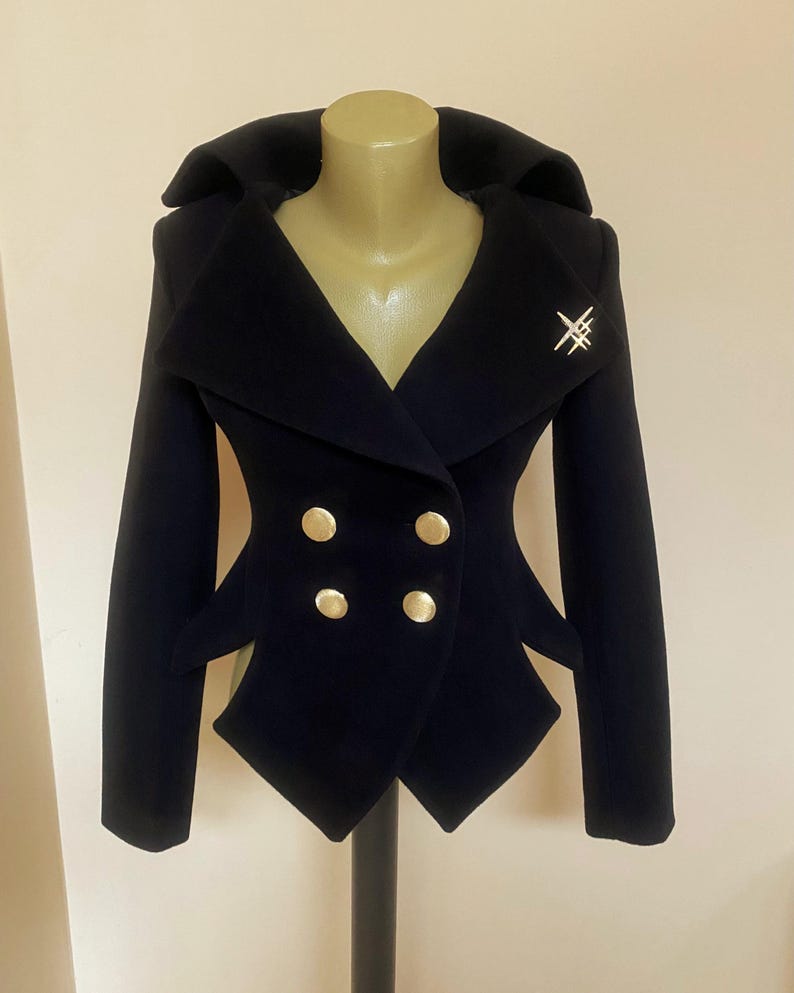 May include: A navy blue double-breasted jacket with gold buttons and a decorative silver pin. The jacket has a unique, tailored design with a wide collar and structured silhouette. The jacket is displayed on a mannequin.