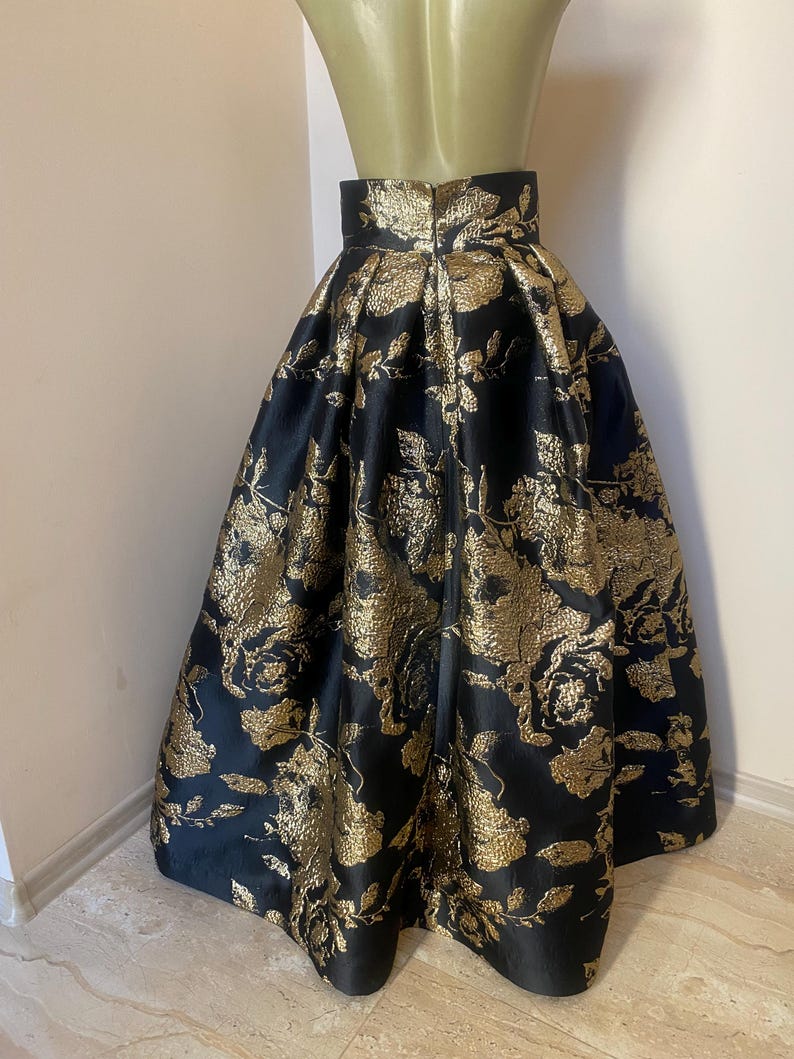 May include: A black and gold floral print skirt. The skirt is made of a shiny fabric and features a high waist and a full, flowing silhouette. The gold floral pattern is detailed and covers the entire skirt.