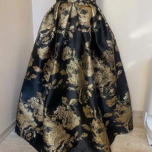 May include: A black and gold floral skirt. The skirt features a high waist and a full, flowing silhouette. The fabric has a textured appearance with a repeating pattern of gold flowers and leaves against a black background. The skirt is displayed on a mannequin.