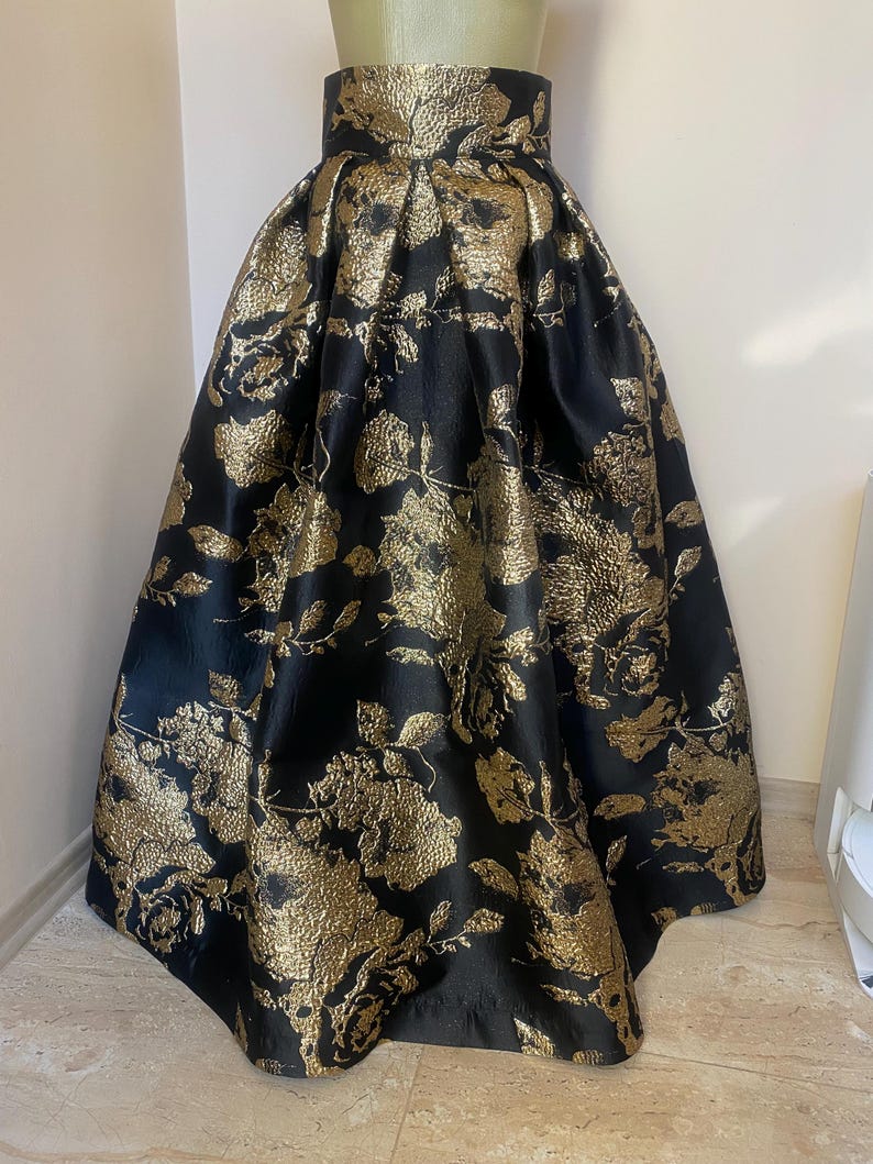 May include: A black and gold floral skirt. The skirt features a high waist and a full, flowing silhouette. The fabric has a textured appearance with a repeating pattern of gold flowers and leaves against a black background. The skirt is displayed on a mannequin.