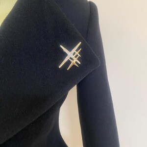 May include: A gold-toned brooch in a starburst design, adorned with clear crystals, is pinned to a dark navy blue coat. The brooch features multiple star-like elements, creating a sparkling accent against the solid fabric. The coat's lapel is visible.