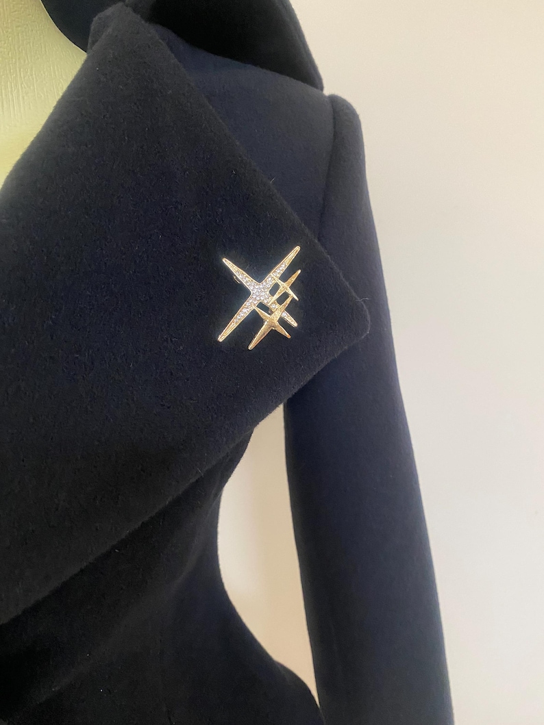 May include: A gold-toned brooch in a starburst design, adorned with clear crystals, is pinned to a dark navy blue coat. The brooch features multiple star-like elements, creating a sparkling accent against the solid fabric. The coat's lapel is visible.