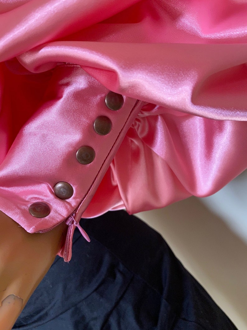 May include: A pink satin blouse with a zipper and button closure on the sleeve. The buttons are silver and the zipper is black.