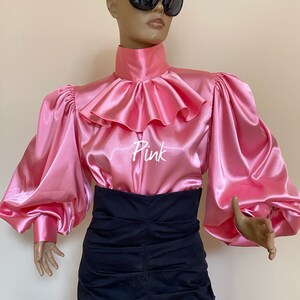 May include: A pink satin blouse with a large ruffled collar and long, puffy sleeves. The blouse has the word "Pink" written on the front.