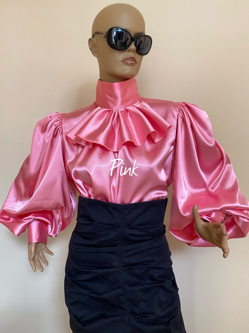 May include: A pink satin blouse with a large ruffled collar and long, puffy sleeves. The blouse has the word "Pink" written on the front.