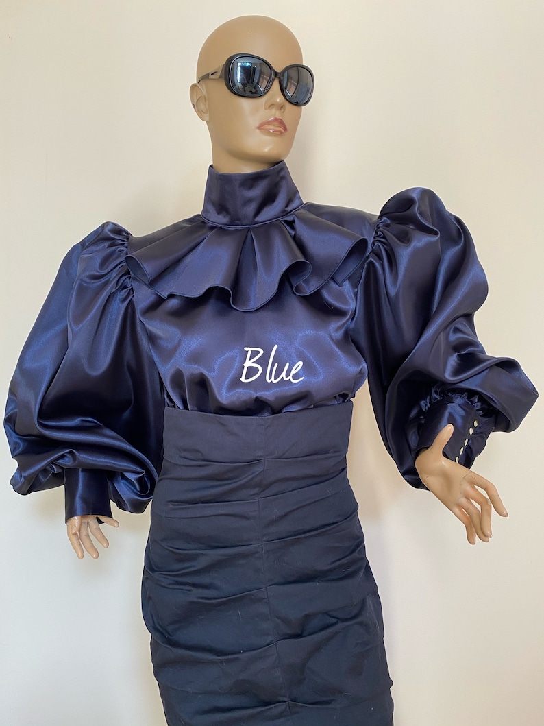 May include: A navy blue satin blouse with a high ruffled collar and large, puffed sleeves. The blouse has the word "Blue" embroidered on the front.