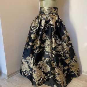 May include: A full, floor-length skirt with a high waist, displayed on a mannequin. The skirt is black with a gold floral pattern. The fabric has a metallic sheen, and the skirt is gathered at the waist, creating a voluminous silhouette. The skirt is suitable for formal events.