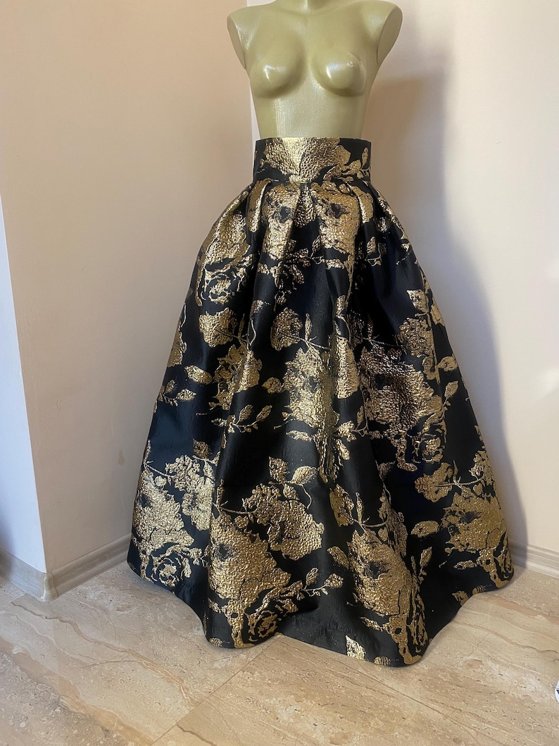 May include: A full, floor-length skirt with a high waist, displayed on a mannequin. The skirt is black with a gold floral pattern. The fabric has a metallic sheen, and the skirt is gathered at the waist, creating a voluminous silhouette. The skirt is suitable for formal events.