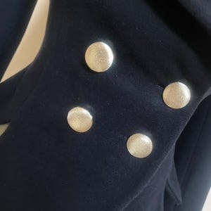 May include: Close-up of a navy blue blazer with four large, gold-colored buttons. The buttons are round and have a textured surface. The blazer's fabric appears to be a soft wool or wool blend.