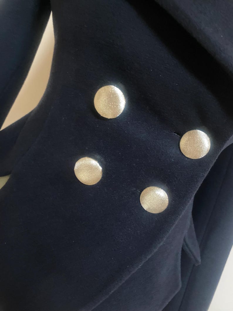 May include: Close-up of a navy blue blazer with four large, gold-colored buttons. The buttons are round and have a textured surface. The blazer's fabric appears to be a soft wool or wool blend.