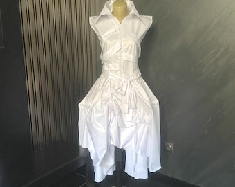 Puede incluir: Un vestido blanco sobre un maniquí. El vestido presenta un cuello camisero, una cremallera frontal y una falda en capas con un dobladillo asimétrico. El vestido está hecho de una tela brillante y crujiente.