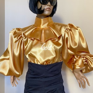 May include: A gold satin blouse with a high ruffled collar and long, puffed sleeves. The blouse has a high neckline and a fitted bodice. The model is wearing a black skirt.