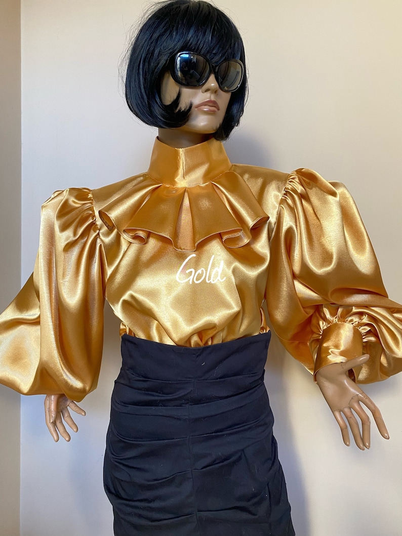 May include: A gold satin blouse with a high ruffled collar and long, puffed sleeves. The blouse has a high neckline and a fitted bodice. The model is wearing a black skirt.