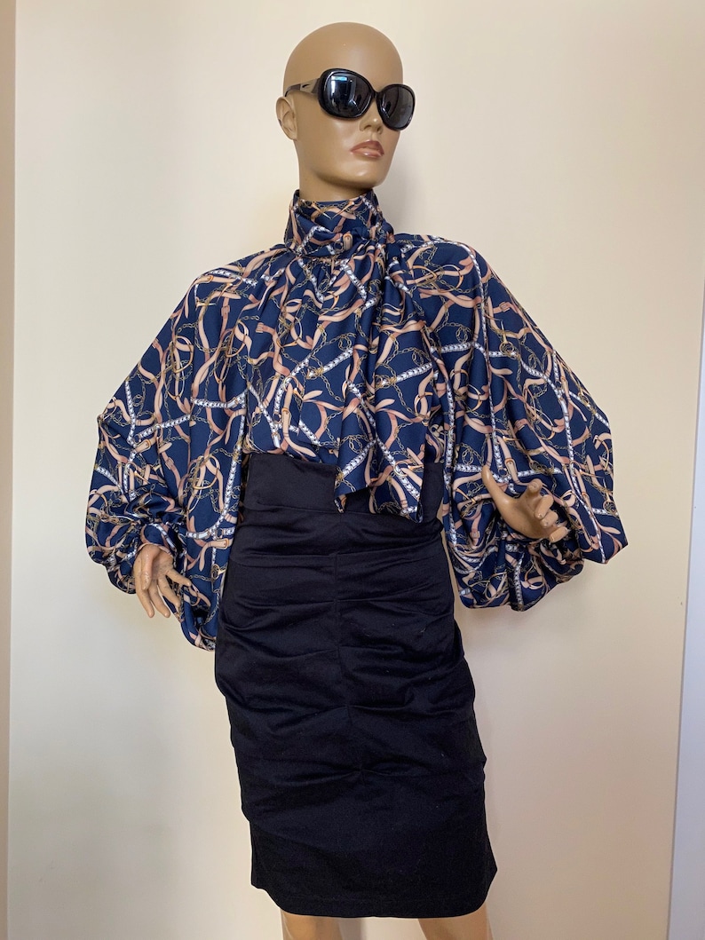 May include: A blue and gold patterned blouse with a high neck and long sleeves. The blouse has a gathered detail at the neck and a loose, flowing fit. The sleeves are gathered at the shoulders and have a wide, bell-shaped cuff.