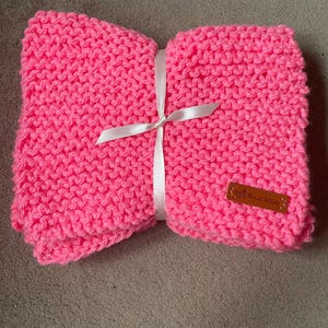 May include: A pink knitted baby blanket with a white ribbon and a brown leather tag that says "Hand Made".