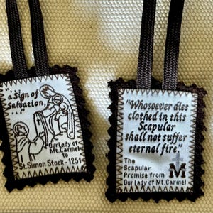 May include: Two brown fabric scapulars with white fabric inserts and brown cords. The left scapular features a black and white image of the Virgin Mary and a child. The text reads "a sign of Salvation... Our Lady of Mt. Carmel to St. Simon Stock - 1251 - ". The right scapular features the text "Whosoever dies clothed in this Scapular shall not suffer eternal fire." The text continues "The Scapular Promise from Our Lady of Mt. Carmel".