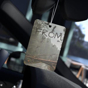 Può includere: Un deodorante per ambienti rettangolare con una scena di foresta e la parola "FROM" scritta a mano. L'immagine raffigura una strada bloccata da alberi caduti, con uccelli in volo. Il deodorante è appeso a un filo.