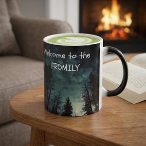 May include: A black and white ceramic mug with the text "Welcome to the FROMILY" against a backdrop of a forest and starry sky. The mug contains a green beverage with latte art and sits on a wooden table.
