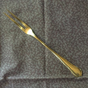 Può includere: Una forchetta dorata a due rebbi con un manico lungo e sottile. La forchetta è appoggiata su uno sfondo di tessuto grigio scuro a fantasia. I rebbi sono affilati e il manico ha un bordo decorativo.