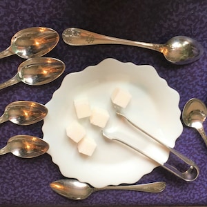 Può includere: Un piatto bianco con cinque cubetti di zucchero e delle pinzette per lo zucchero argentate. Ci sono sette cucchiaini d'argento su uno sfondo viola.
